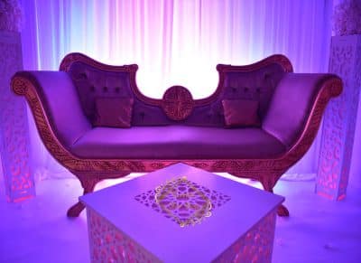 Luxueuse banquette marocaine en velours pourpre et or pour mariage, avec table basse et éclairage magenta ambiant.