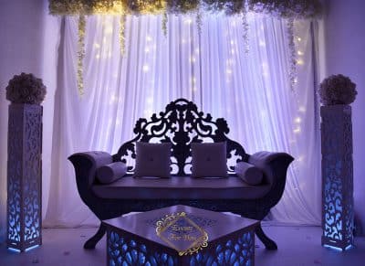 Canapé de mariage noir sculpté, scène événementielle romantique. Piliers et table avec éclairage bleu. Rideaux et guirlandes lumineuses. Logo : Events For You.