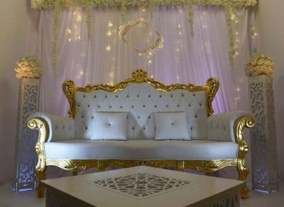 Canapé des mariés blanc capitonné et doré, avec coussins. Décor de scène luxueux : voilages, éclairages LED, colonnes florales.