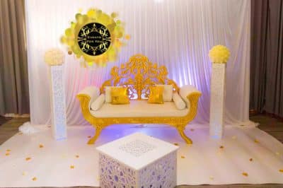 Canapé royal doré, fond de scène drapé blanc éclairé, colonnes florales jaunes et table cubique ajourée. Décor luxe "Events For You".