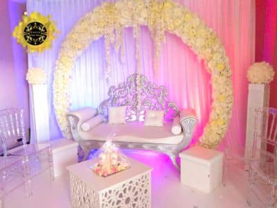 Canapé baroque argenté luxueux sur scène de mariage. Grande arche florale blanche, tables ajourées, chaises acryliques et éclairage d'ambiance violet/rose.