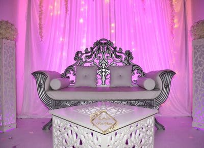 Canapé trône argenté pour événement sur scène éclairée en rose fuchsia. Rideaux blancs drapés, table sculptée. Panneau : Events For You.