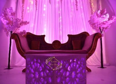 Canapé de réception pourpre antique, flanqué d'arbres en fleurs de cerisier. Table lumineuse ornée de motifs, sous un éclairage magenta. Events For You.