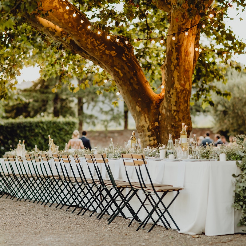 Dîner de Mariage Extérieur | Décor sous Arbre et Lumières Longue table de réception extérieure dressée pour un mariage sous un platane éclairé par des guirlandes. Chaises bistrot rustiques.