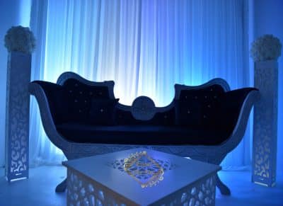 Décor lounge de mariage luxueux avec sofa baroque en velours noir et structure blanche sculptée. Lumière bleue intense, colonnes et table ajourées.