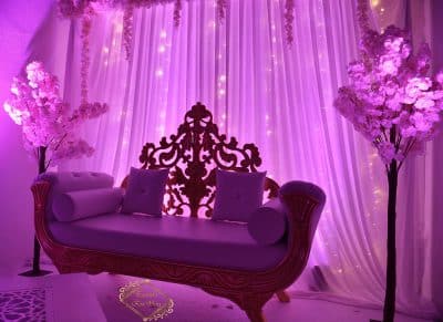 Décoration de mariage violette : élégant sofa orné (dossier sculpté or) flanqué d'arbres en fleurs roses. Rideaux lumineux.