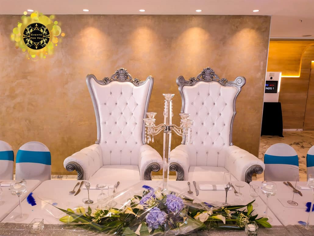 Deux trônes d'honneur blancs royaux pour mariage, capitonnés et bordés d'argent, devant un mur texturé doré. Table décorée de fleurs, candélabre. Logo Events For You.