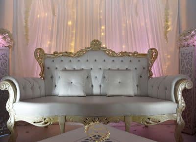 Élégant trône de mariage blanc et doré, capitonné, sur une scène décorée de voilages illuminés pour un événement de luxe.