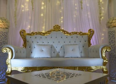 Trône de mariés baroque blanc et or, avec capitonnage et coussins, sur une scène décorée de voilages lumineux et de colonnes fleuries.