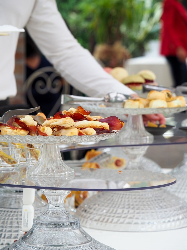 Apéritifs et buffet élégant sur plateau en verre ciselé Un plateau en verre ciselé rempli de feuilletés au jambon cru et salés sur un buffet de réception. Un serveur tend la main derrière la verrerie.