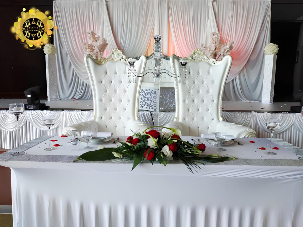 Fauteuils trône blancs : table d'honneur mariage de luxe. Deux fauteuils trône blancs capitonnés sur la table d'honneur d'un mariage. Décoration luxueuse, drapé blanc, roses rouges et logo "EVENTS FOR YOU".